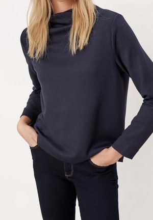 Femme aux longs cheveux blonds portant un haut à manches longues bleu marine foncé et un jean foncé, les mains dans les poches, debout devant un fond uni.