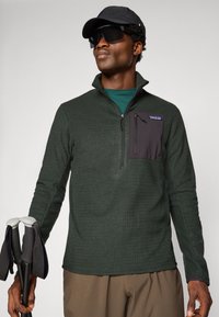 Pull-over vert foncé en tissu texturé, avec une poche poitrine noire à fermeture éclair, associé à des bâtons de randonnée et une casquette noire.