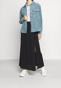 Camicia in velluto a coste color teal con due tasche sul petto, abbinata a una gonna maxi nera con uno spacco laterale e sneaker bianche oversized.