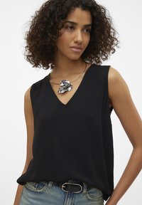 Black sleeveless top with a V-neck and textured fabric. Worn with light blue denim jeans and a metallic floral necklace as an accent.