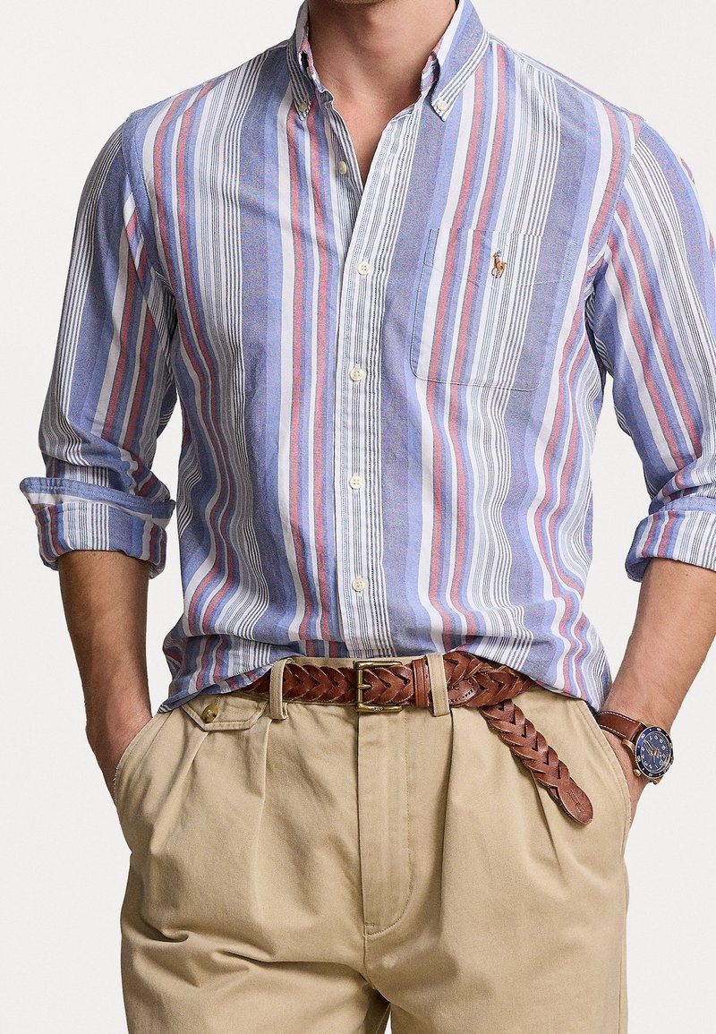 Camisa de botones a rayas en azul, rosa y blanco, con un bolsillo en el pecho. Combinada con pantalones caqui y un cinturón de cuero trenzado marrón.