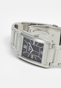 Relógio de pulso prateado com mostrador retangular, mostrador preto, números romanos brancos e bracelete de metal prateado. Apresenta uma coroa de lado.