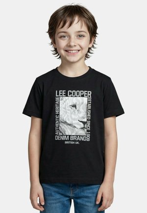 Enfant souriant aux cheveux courts portant un t-shirt noir Lee Cooper avec un graphisme de lion et un jean bleu sur un fond blanc uni.