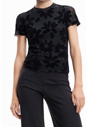 Frau trägt ein schwarzes, durchsichtiges Kurzarm-Top mit großen Blumenmustern und schwarze Hose, steht vor einem weißen Hintergrund.