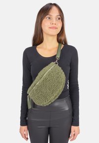 Sac bandoulière en fausse fourrure vert olive avec une forme arrondie, fermeture éclair argentée et surface texturée. Sangle attachée en matériau vert assorti.