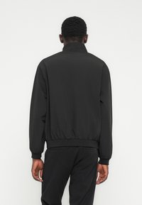 Μαύρο ελαφρύ μπουφάν με ψηλό γιακά, ελαστικά μανίκια και ζώνη στη μέση. Λείο υλικό, μινιμαλιστικός σχεδιασμός και χωρίς εμφανές λογότυπο.