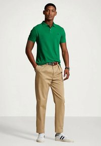 Άνδρας που φορά πράσινο πόλο μπλουζάκι, μπεζ παντελόνι με ζώνη, λευκά αθλητικά παπούτσια, στέκει με τα χέρια στις τσέπες του σε απλή φόντο.