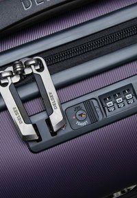 Maleta rígida de color púrpura de Delsey con tiradores de cremallera duales, cerradura de combinación y superficie texturizada con líneas verticales.