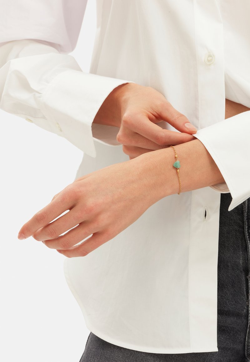 Bracciale in oro con un accento a cuore di turchese, indossato su un polso, con una camicia bianca a maniche lunghe sullo sfondo.
