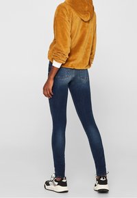 Sudadera de pana amarilla con capucha, combinada con jeans ajustados de color azul oscuro y zapatillas negras y blancas, vista desde atrás.