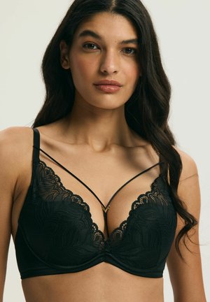 Kobieta z długimi ciemnymi włosami, ubrana w czarny koronkowy biustonosz z ozdobnymi paskami, patrząca prosto w kamerę na jednolitym tle.