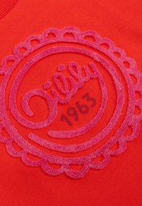 Rode sweatshirt met een gestructureerd roze circulair logo met gekartelde randen, het woord "Lilly" en "1963" in het midden.