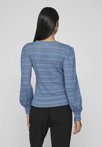 Modrá pletená svetr s texturou pruhovaného vzoru, nafouknutými rukávy a přiléhavým pasem. Nosený s černými kalhotami.