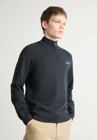 Sudadera de cuello con cremallera negra en tejido de punto texturizado, con mangas largas y un sutil logo en el pecho. Ajuste clásico y textura suave.