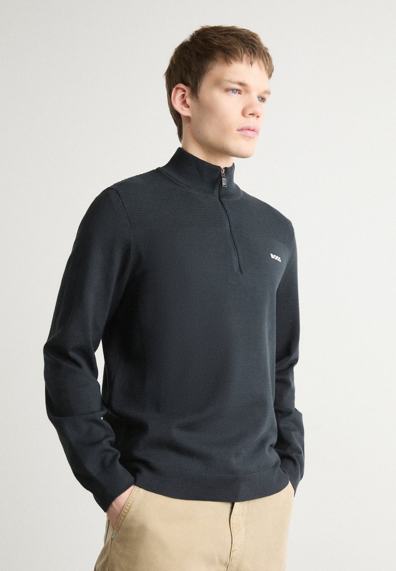 Sudadera de cuello con cremallera negra en tejido de punto texturizado, con mangas largas y un sutil logo en el pecho. Ajuste clásico y textura suave.