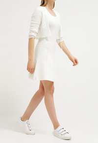 Bílý pletený svetr s krátkým střihem a hvězdicovým vzorem, v kombinaci s upnutými bílými šaty. Modelka má na sobě bílé slip-on tenisky.