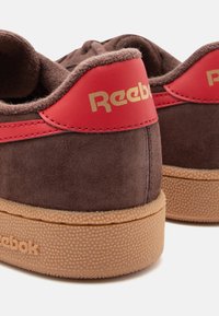 Brune semskede sneakers med røde læderdetaljer, guld "Reebok"-logo og tekstureret gummiydersål. Afrundet form og polstret krave.