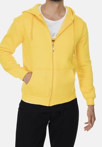 Uomo che indossa un hoodie giallo con zip e tasche frontali sopra una maglietta bianca e pantaloni neri, con una mano in tasca, visibile dalla testa ai fianchi.
