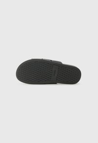 Black rubber sandal sole with textured grip pattern, featuring a flat design and a logo imprint near the heel. Smooth surface, minimal detailing.