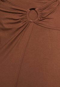 Brun tygblus med en samlad design och en central cirkulär acccent, slät textur och vertikala sömdetaljer.