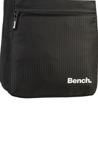 Schwarze Tasche aus strapazierfähigem Gewebe mit einem strukturierten, hexagonalen Muster auf der Vorderseite und einer Reißverschlusstasche. Markenlogo in Weiß.