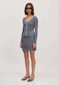 Grauer, gerippter Cardigan mit tiefem V-Ausschnitt und weißen Akzenten, kombiniert mit einem passenden grauen Minirock mit zwei Seitentaschen. Schwarze Sandalen.