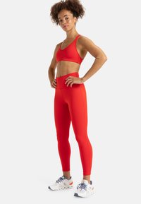 Soutien-gorge de sport rouge et leggings taille haute assortis en tissu lisse et extensible. Associés à des chaussures de sport blanches avec des accents bleus et oranges.