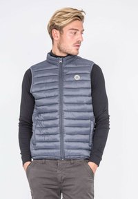 Veste sans manches grise avec un col montant, fermeture éclair à l'avant et design matelassé horizontal. Présente une applique de logo et est portée sur une chemise noire à manches longues.