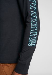 Camisa negra de manga larga con texto vertical azul claro "WRGHIW" y pequeños diseños cuadrados azules, combinada con pantalones marrones.