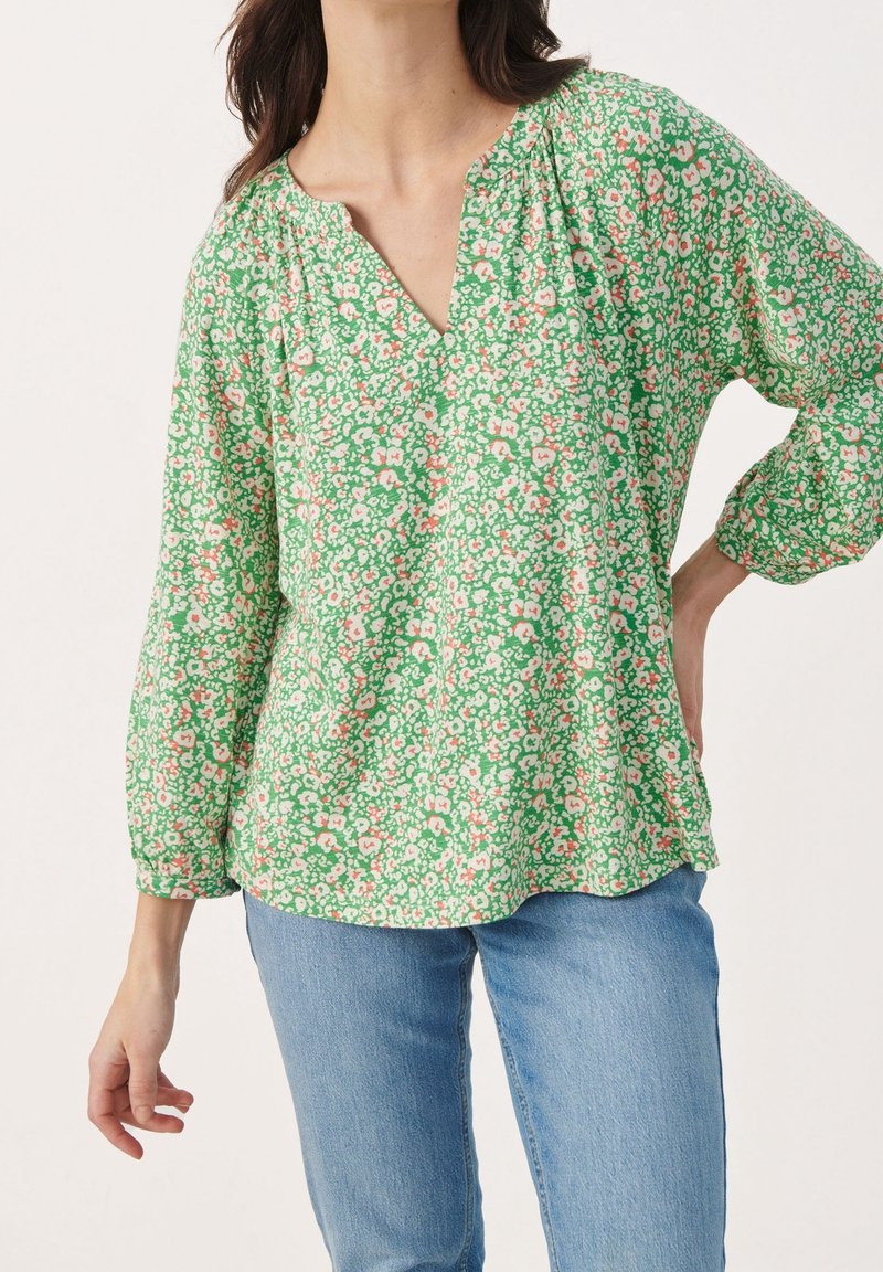 Femme portant un chemisier vert à fleurs à manches longues avec une encolure fendue, associé à un jean bleu clair, posant avec une main sur la hanche.