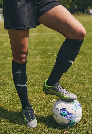 Jugador de fútbol con pantalones cortos y calcetas negras controla un balón blanco y azul con un pie en un campo de césped.