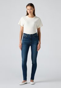 Svetle krémové tričko s krátkým rukávem v kombinaci s tmavě modrými skinny džíny a bílými slip-on botami, s klasickým střihem a minimálními detaily.