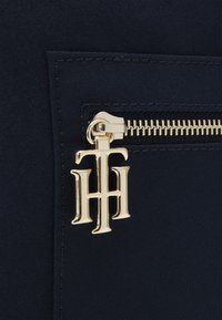 Bolso de tela azul marino con cremallera dorada y un charm con el logotipo. La textura es suave, con un diseño sólido y acentos de costura visibles.