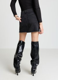 Mini-jupe en denim noir avec un ourlet effiloché et des poches arrière, associée à des bottes en cuir noir brillant montant jusqu'aux genoux avec des talons pointus.