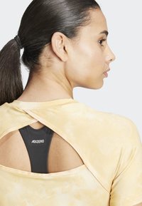 Maglietta atletica gialla tie-dye con schiena a taglio, che rivela un reggiseno sportivo nero con il marchio "ADIZERO". Tessuto liscio, design aderente.