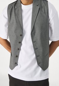 Gilet grigio a fantasia con vestibilità sartoriale, sei bottoni, rever a punta e due tasche, indossato sopra una t-shirt bianca.