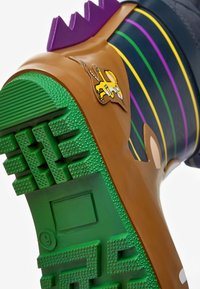 Botte en caoutchouc marron avec une semelle texturée verte, des rainures violettes en haut et une patch d'animal jaune. Présente des détails rayés en bleu, vert et violet.