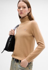 Sweter typu golf z miękkiej dzianiny w odcieniu camel, z małym logo na piersi, w zestawieniu z dżinsami o wysokim stanie i czarną torbą.