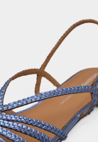 Κοντινό πλάνο καφέ πλατφόρμας σανδάλι με μεταλλικά μπλε πλεκτά λουριά και βρόχο για τον αστράγαλο σε λευκό φόντο.