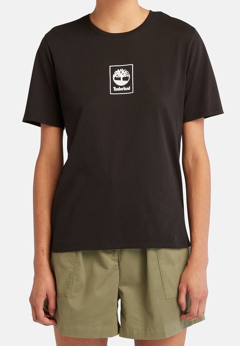 Personne portant un t-shirt Timberland noir à manches courtes et un short vert olive, debout, les bras détendus le long du corps.