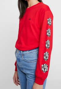 Röd långärmad skjorta med svart "OBEY"-logo på bröstet och grafiska detaljer på ärmarna i ett djärvt, kontrasterande mönster. Tillverkad av bomull.