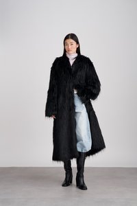 Eine Frau trägt einen langen schwarzen Kunstfellmantel, helle blaue Jeans, schwarze Absatzstiefel und einen lavendelfarbenen Rollkragenpullover und steht auf einem grauen Boden.