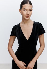 Abito in velluto nero con scollo profondo a V, maniche corte a sbuffo e corpino aderente. Tessuto liscio e silhouette elegante, su sfondo semplice.