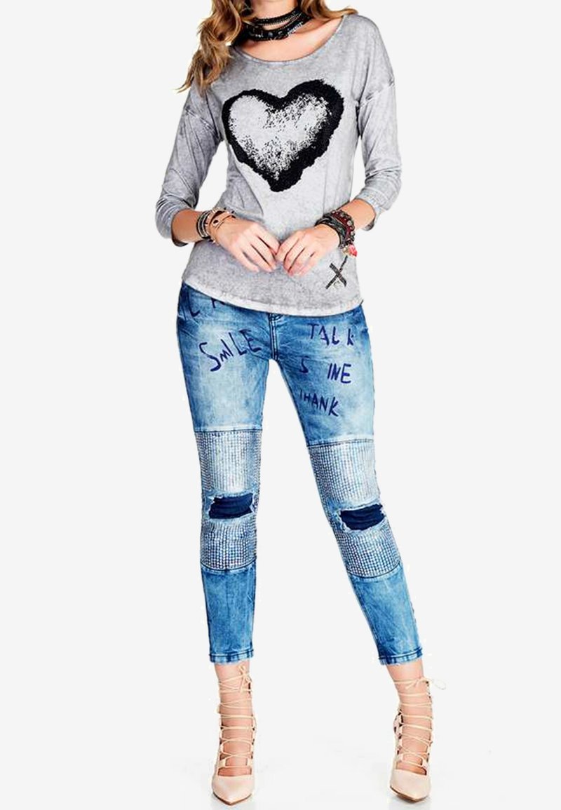 Femme portant un chemisier gris à manches longues avec un grand cœur noir et un jean bleu orné de texte, de coudières côtelées, et des talons beiges à lacets.