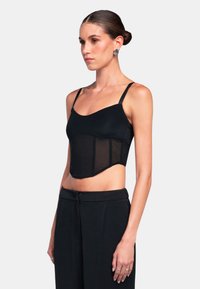 Czarny crop top wykonany z gładkiego materiału z przezroczystymi panelami, regulowanymi ramiączkami i usztywnioną konstrukcją. W połączeniu z czarnymi spodniami o wysokim stanie.