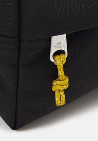 Esquina de una bolsa de tela negra con un tirador de cremallera plateado grabado "The North Face" y un cordón anudado de color amarillo con puntos blancos adjunto.