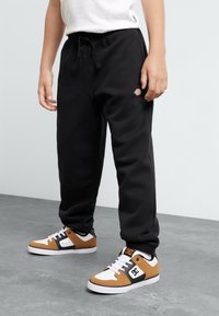 Black sweatpants with elastic waistband and logo detail on the hip, paired with brown, white, and black sneakers with contrasting accents.