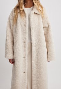 Cremefarvet pelsfrakke med blødt, struktureret stof, krave og knaplukning ned foran. Lange ærmer og knælangt design.