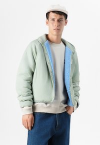Lichtgroene bomberjack met een blauwe fleece voering, drukknoopsluiting en ribgebreide manchetten, gelaagd over een beige sweatshirt en denim jeans.
