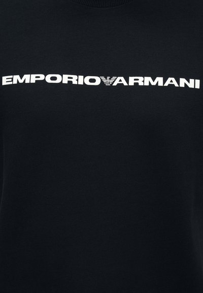 Μαύρο ύφασμα με κεντρικό λευκό κείμενο λογότυπου Emporio Armani που περιλαμβάνει το έμβλημα αετού της μάρκας ανάμεσα στις λέξεις.